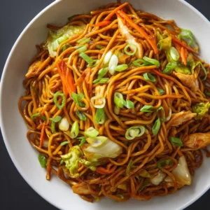 A close-up overhead shot of chow mein noodles being stir-fried in a wok with chicken and fresh vegetables like carrots and cabbage.