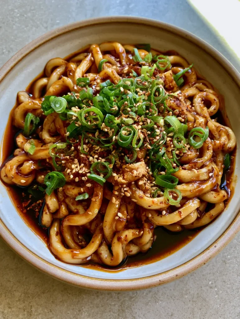 Close-up of chewy udon noodles tangled in a viscous brown Udon Noodle Sauce with visible scallions and chili flakes.